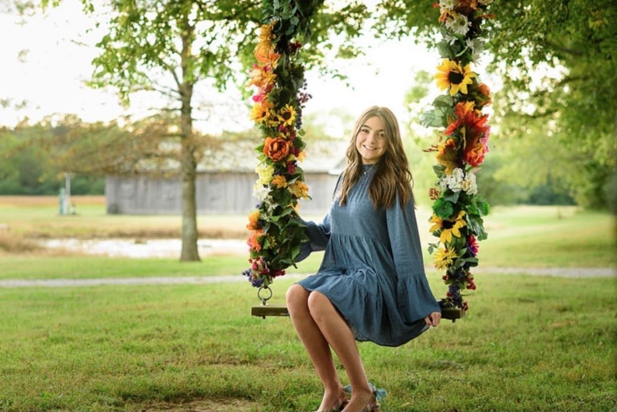 Young girl smiling on a floral-decorated swing outdoors, surrounded by lush green trees in a sunny park setting.