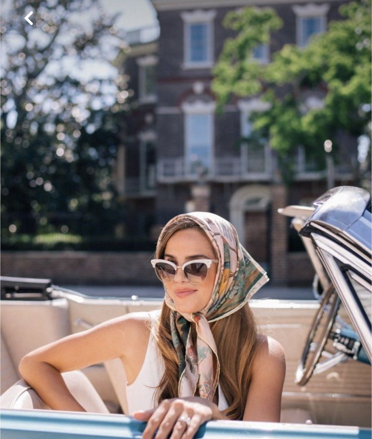 Woman in vintage scarf and cat-eye sunglasses sitting in a classic convertible car on a sunny day.