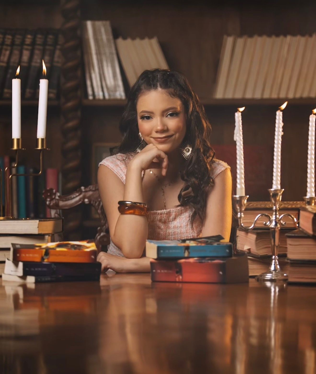Young woman smiling at a wooden desk surrounded by stacked books and lit candles in a cozy library setting.