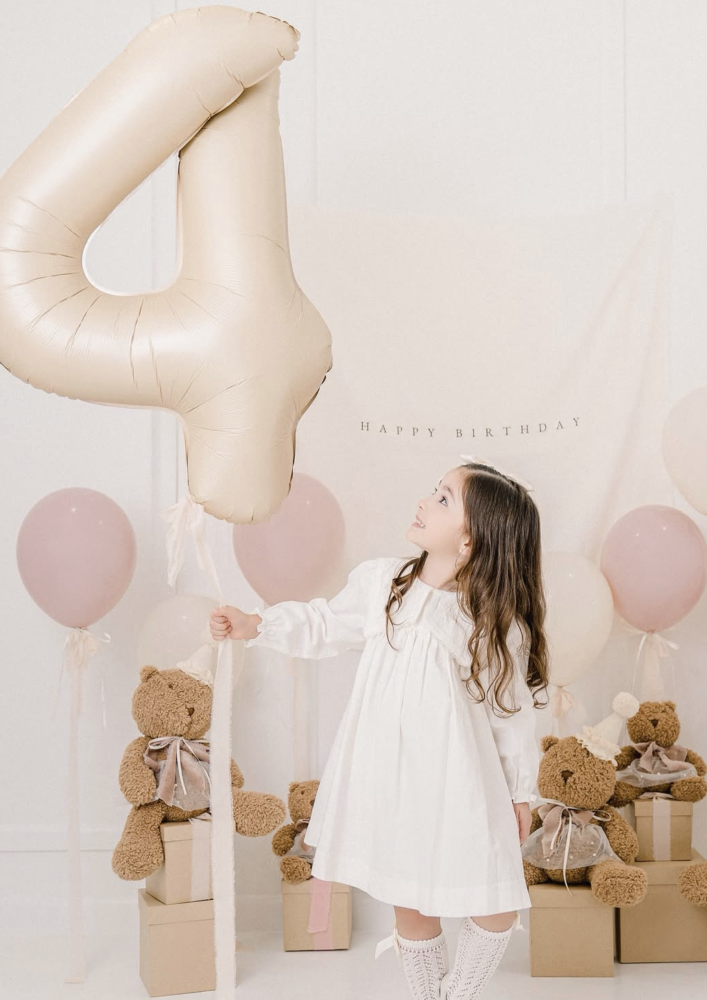 A young girl in a white dress celebrates her 4th birthday with teddy bears, pink balloons, and a large number 4 balloon.