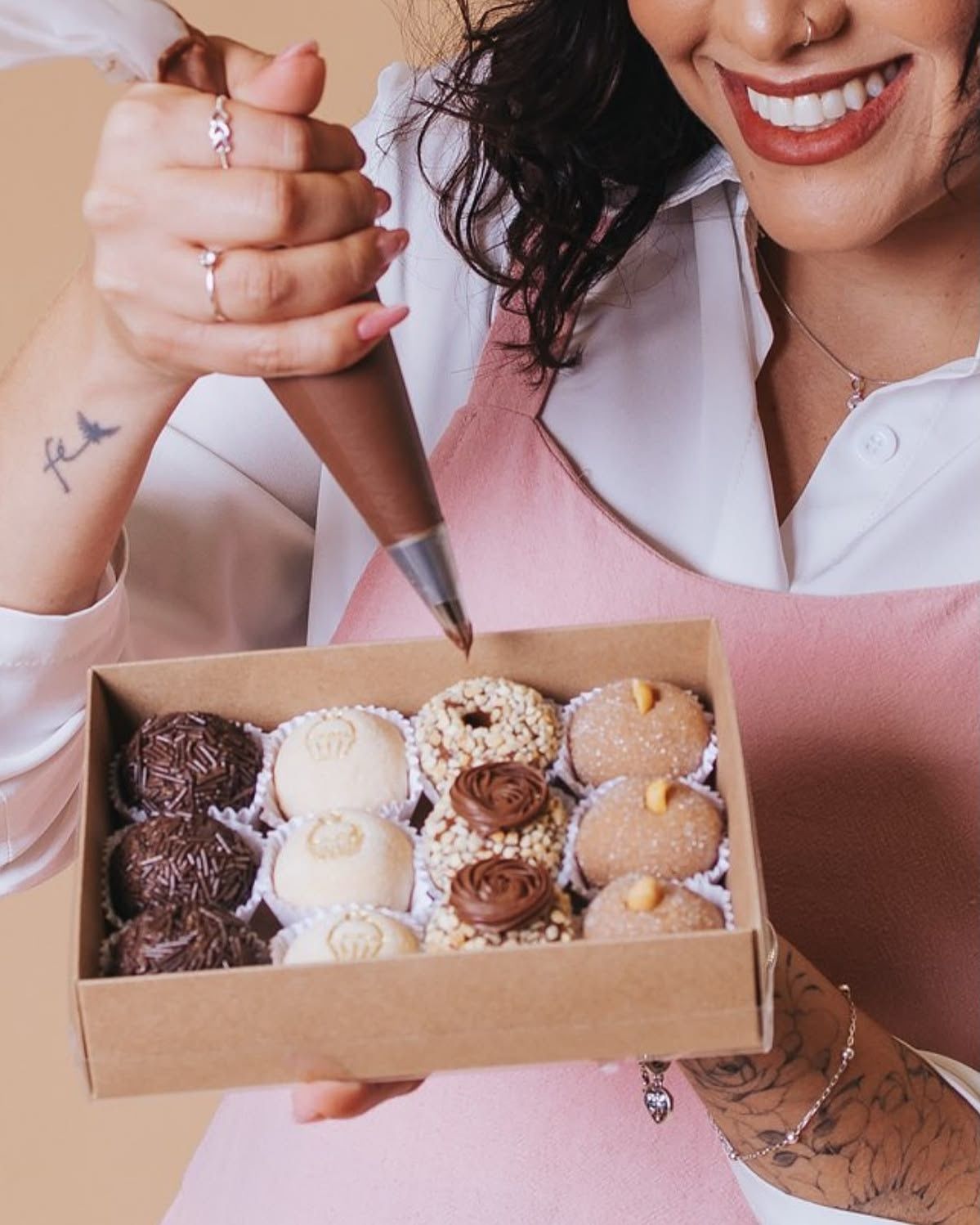 Smiling woman in pink apron holding a box of assorted chocolate truffles while piping frosting.