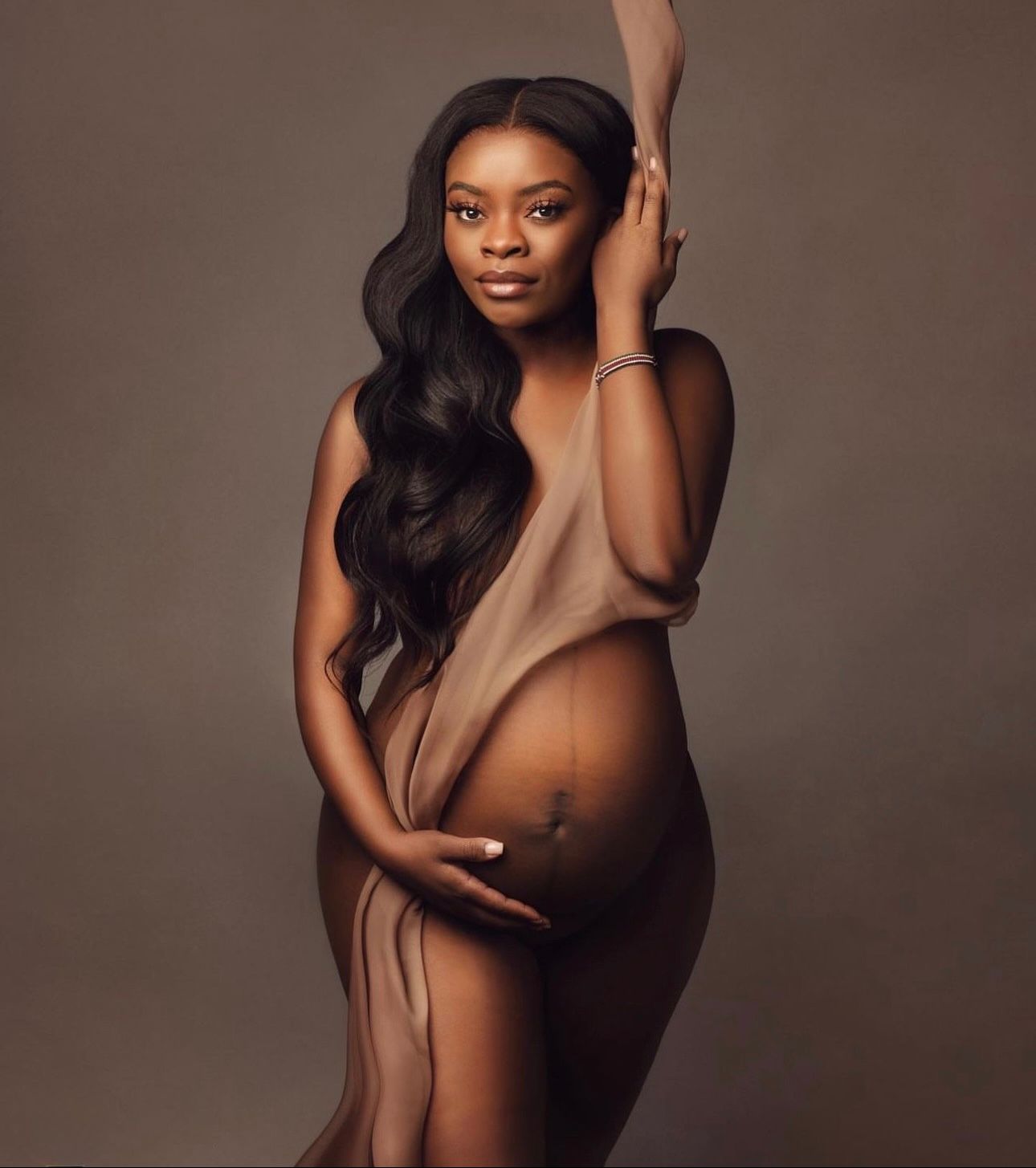 Pregnant woman posing elegantly draped in sheer fabric, cradling baby bump in a professional maternity photo shoot.