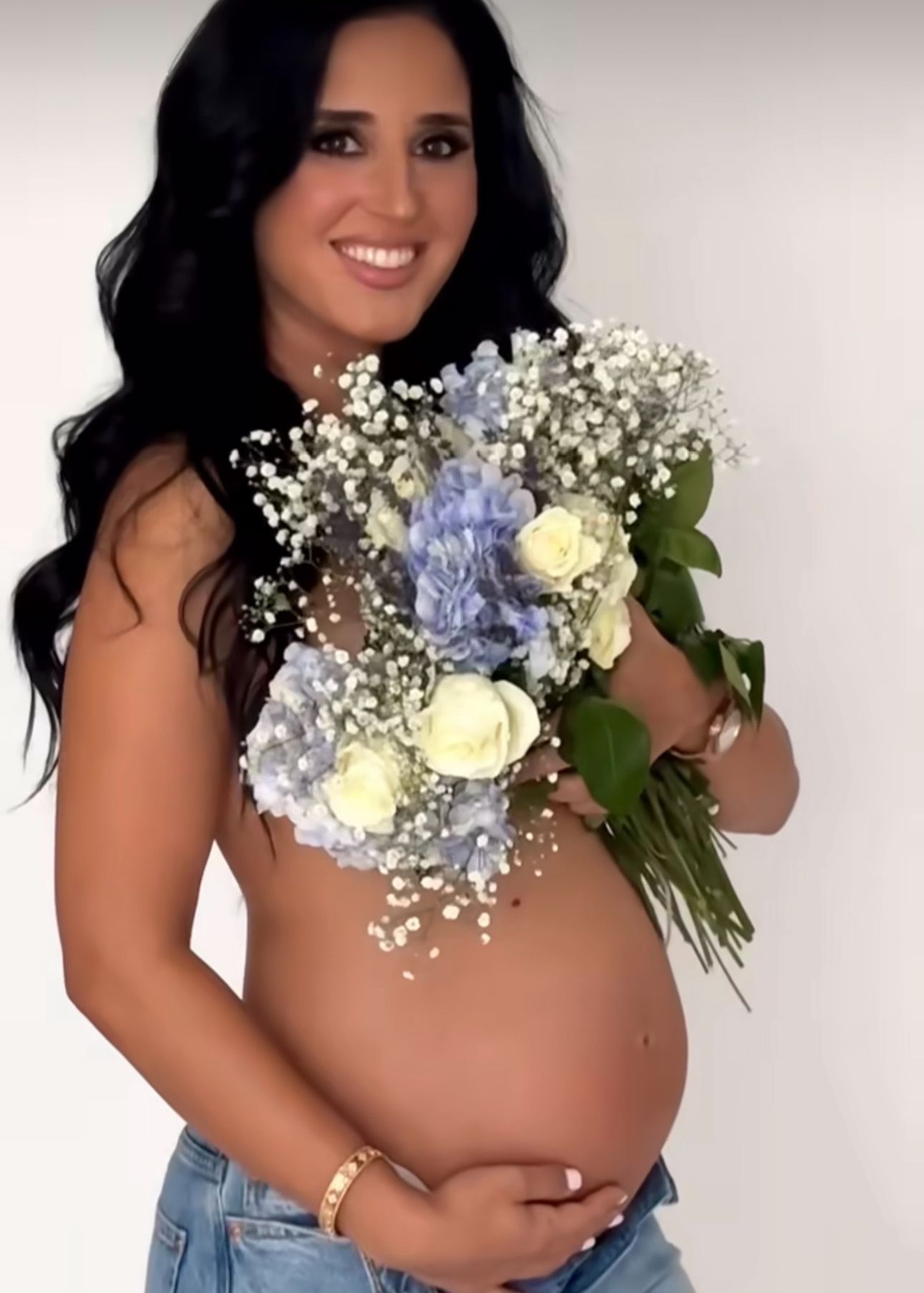 Pregnant woman holding blue hydrangeas and white roses bouquet, wearing jeans, smiling against white background.