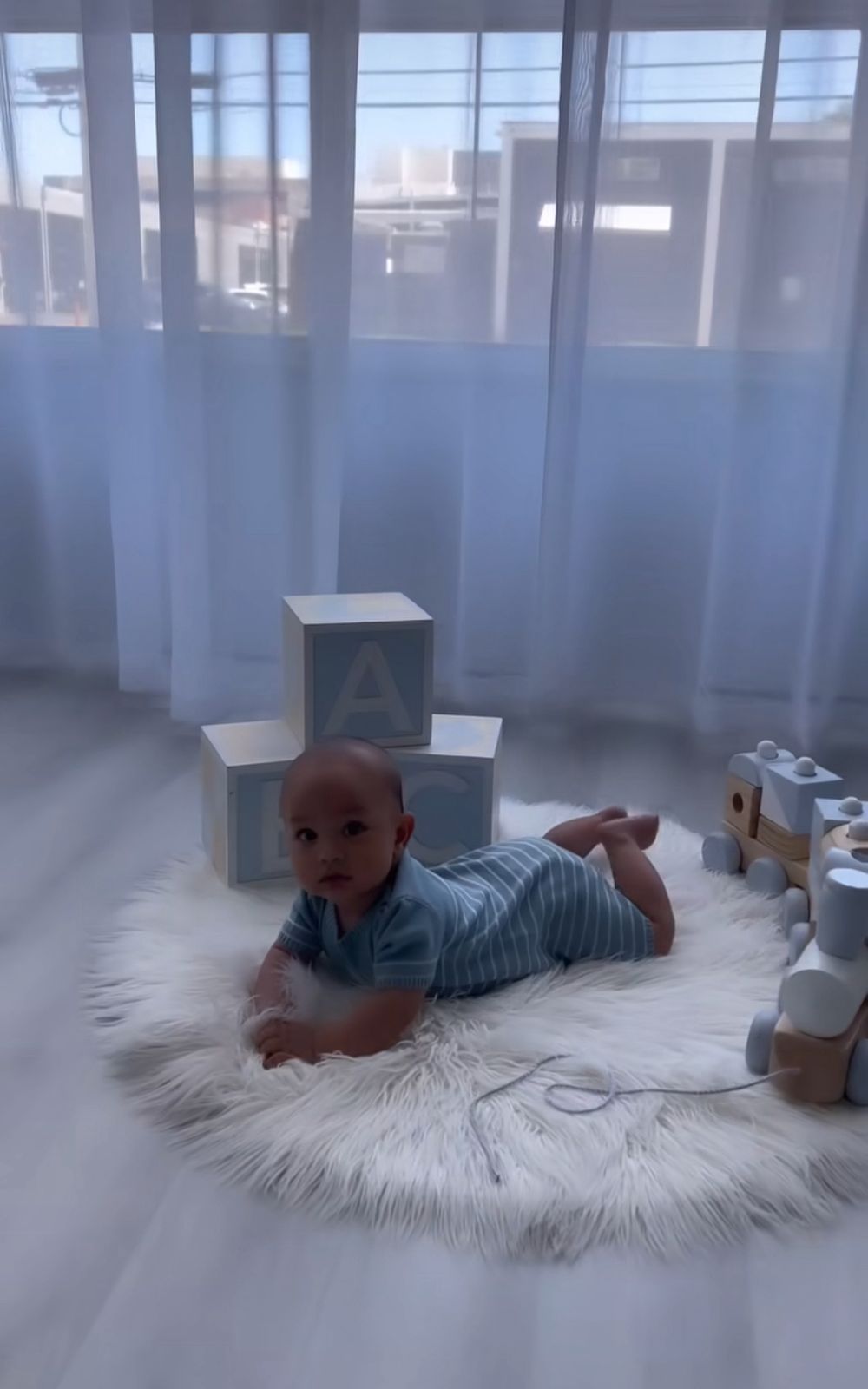 A baby lies on a fluffy white rug in a room with blue curtains and natural light streaming through the windows.