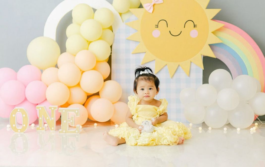 A pastel yellow and pink party setup with balloons, rainbow decorations, and a sun-themed backdrop for a celebration.
