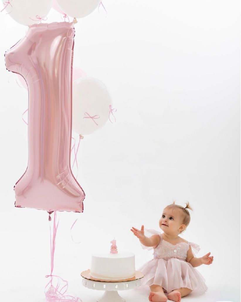 Baby in white dress sits next to birthday cake with large pink number 1 balloon for first birthday celebration.
