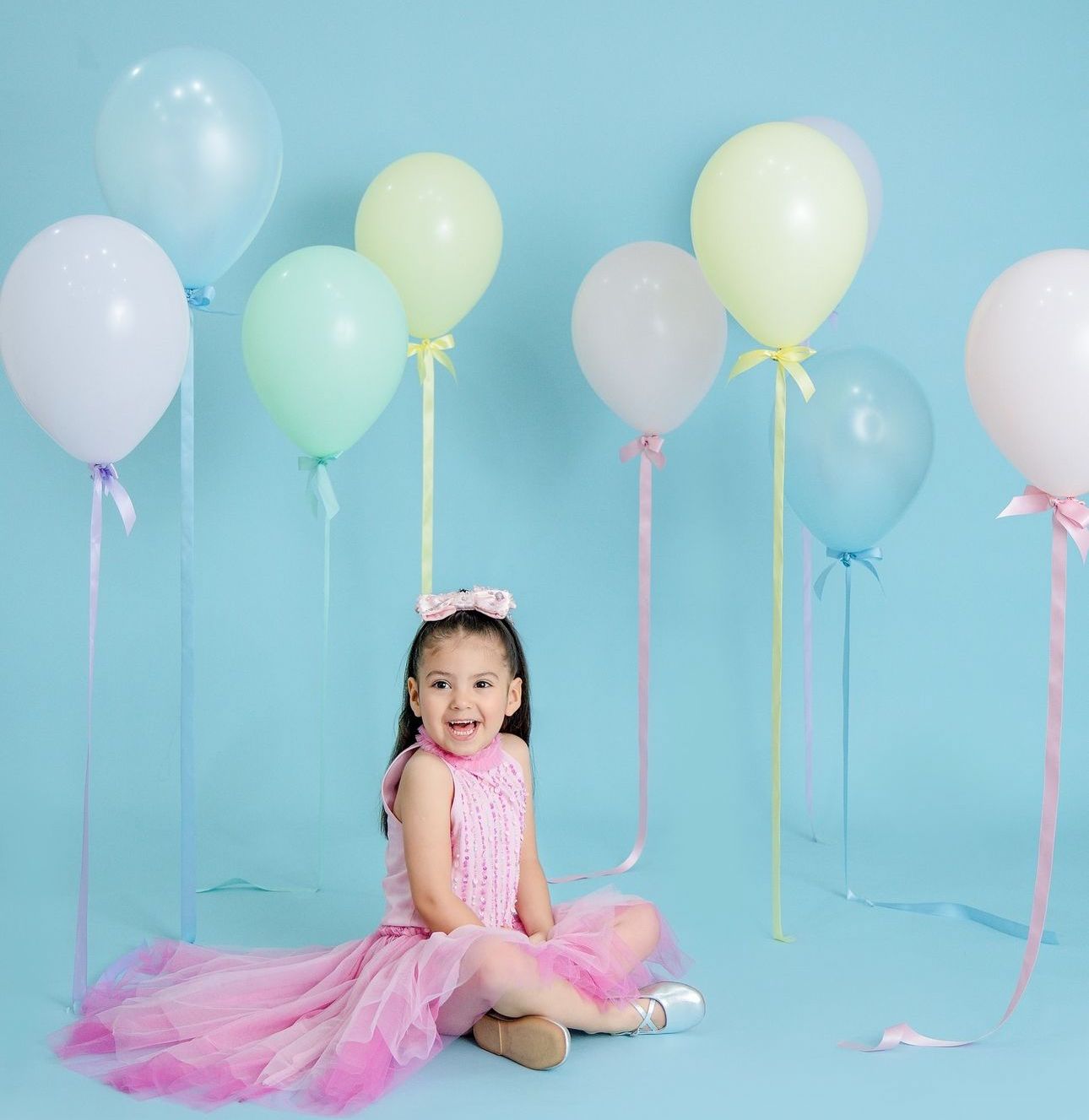 A child in a pink tulle dress sits on the floor surrounded by pastel balloons against a light blue backdrop.