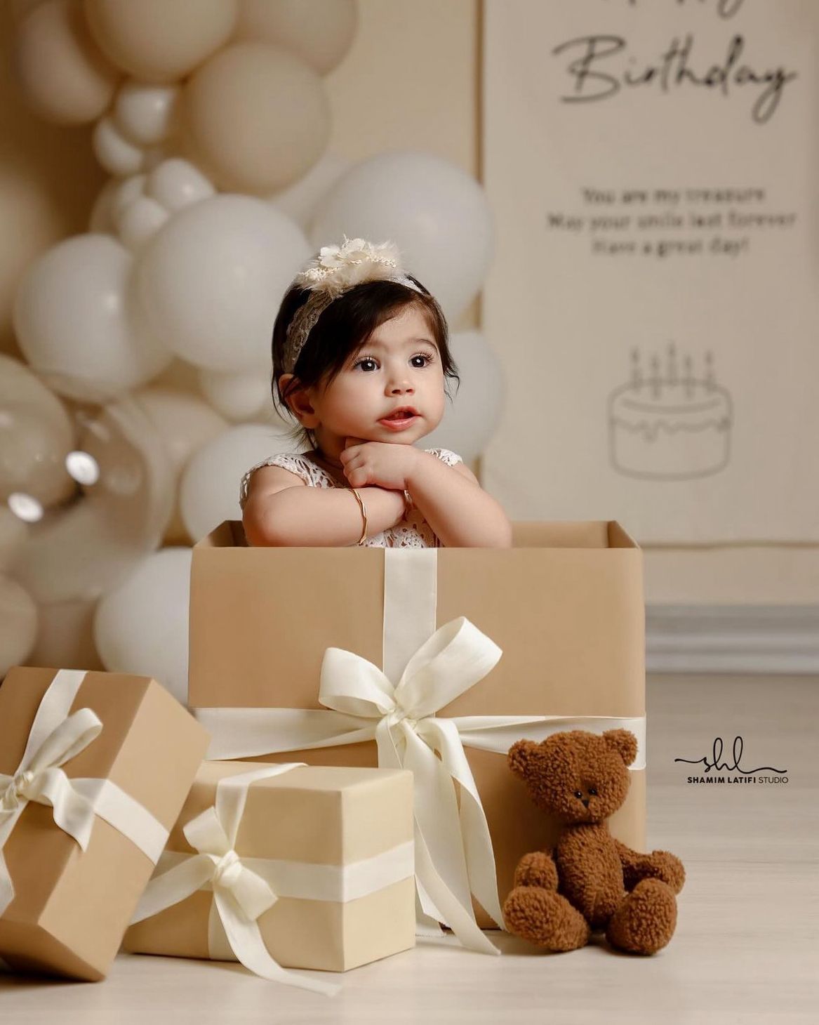 A baby sits in a cardboard box surrounded by wrapped presents, balloons, and a teddy bear for a birthday photoshoot.