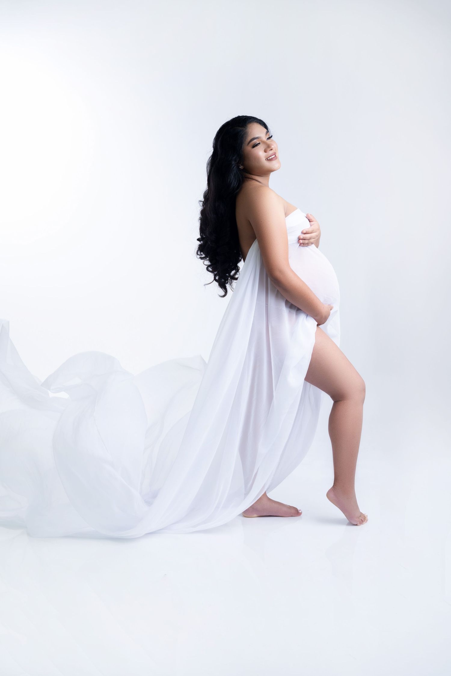 Graceful maternity silhouette in white flowing fabric against light background.