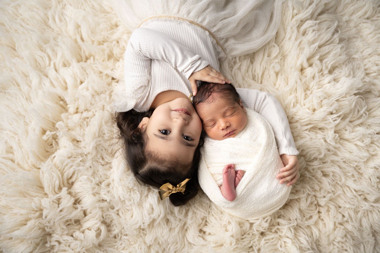 Two siblings lying on cream fluffy rug during professional newborn photo session.