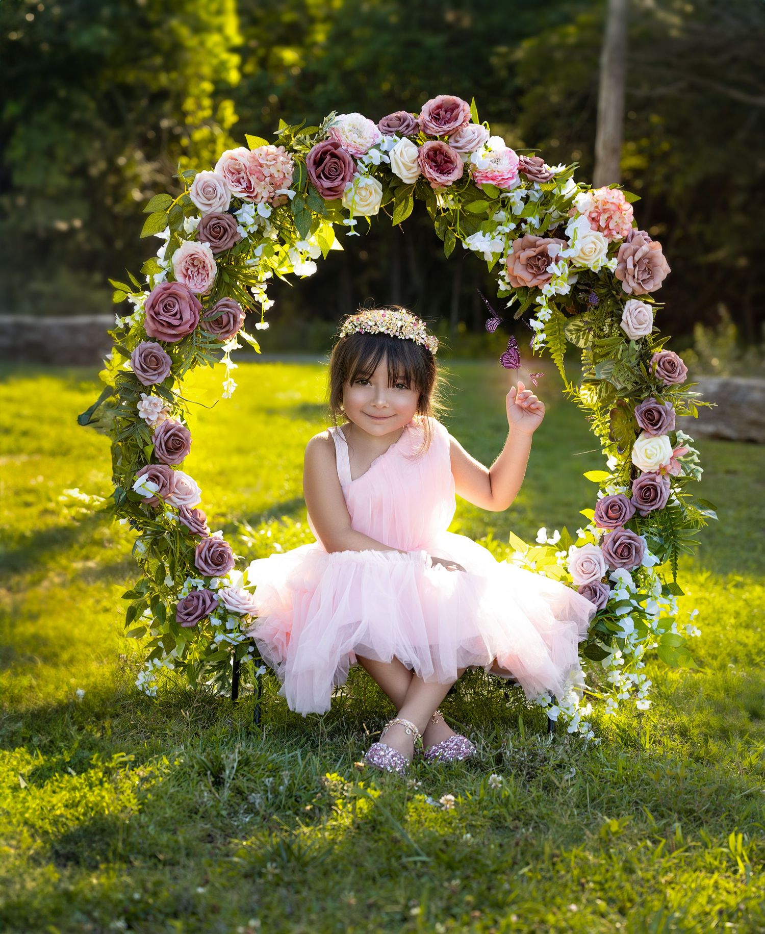 Floral arch with pink and purple roses frames pink tulle dress in sunny garden setting.