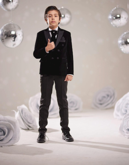 Young boy in black formal suit poses in studio setting with silver disco balls and white rose backdrop.