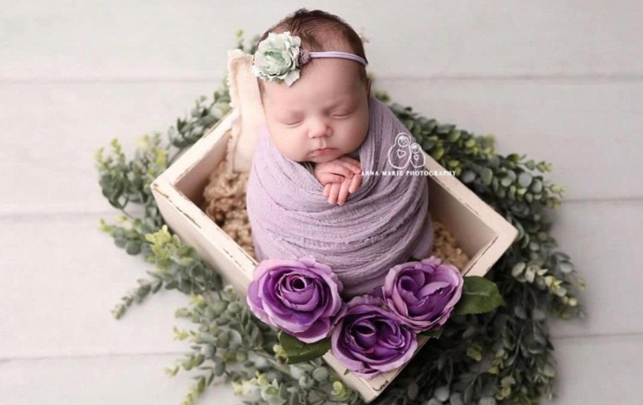 Sleeping newborn wrapped in purple swaddle lying in wooden box with purple flowers and greenery decorations.