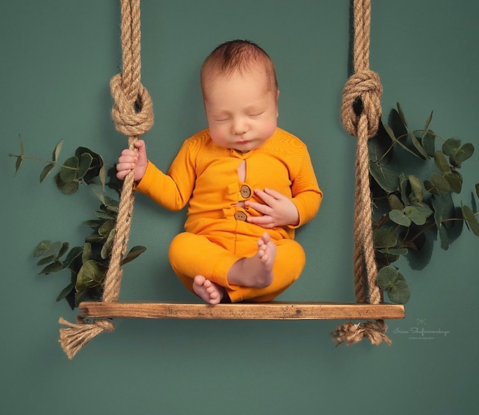 A small child in bright orange clothing sits on a wooden rope swing against a teal background with greenery.