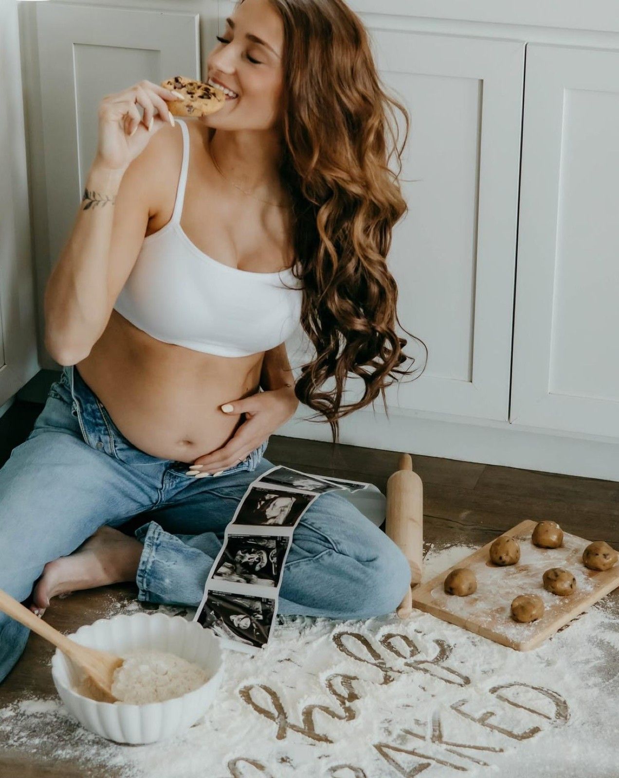 Pregnant woman sitting on flour-dusted floor with ultrasound photos, cookie dough, and rolling pin, eating a cookie.