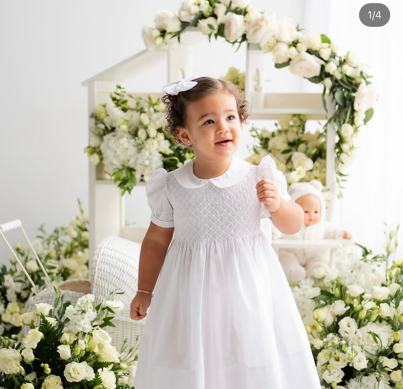A toddler girl smiles in a white smocked dress with peter pan collar surrounded by white floral arrangements.