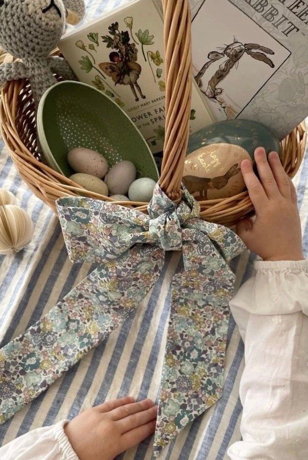 Hands arranging floral fabric and blue striped textiles on a white surface with woven baskets and decorative eggs.