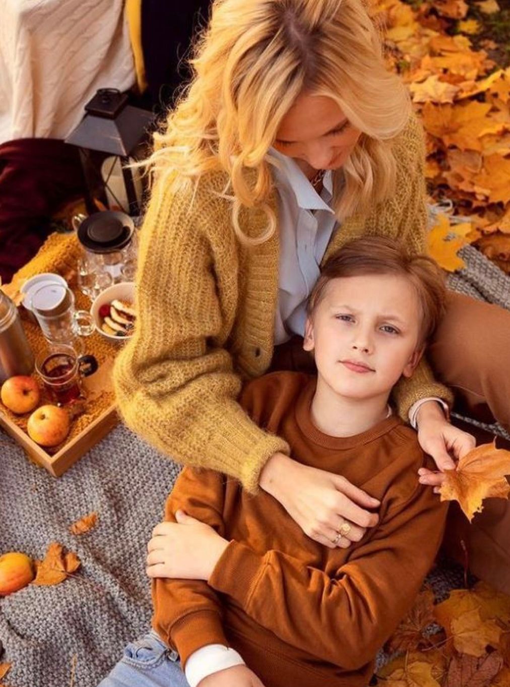 A child in a brown sweater lies on fallen autumn leaves with autumn-themed decor and pumpkins in warm lighting.