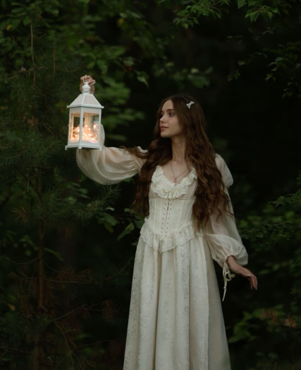 A woman in a white Victorian dress holds a glowing lantern in a dark forest.