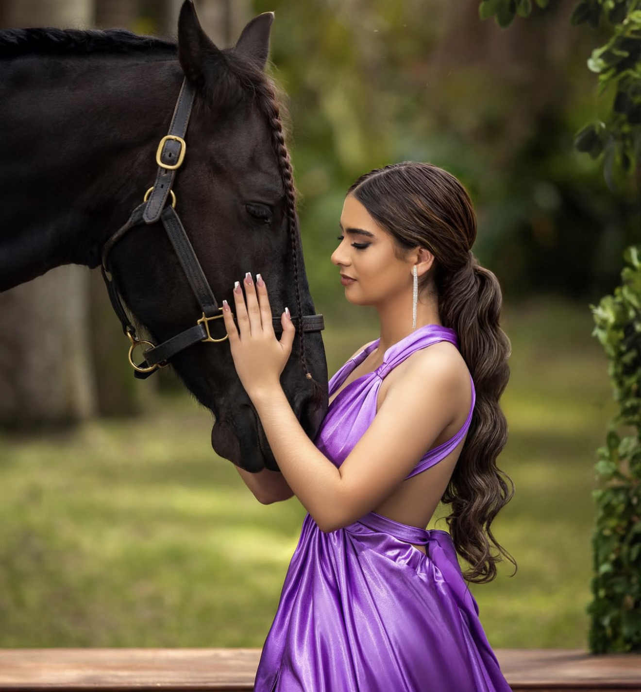 Young woman in purple satin gown gently touching a black horse outdoors in a lush green setting.