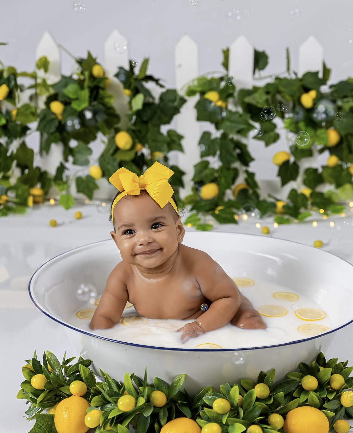 Smiling baby in yellow bow sits in milk bath with lemons, surrounded by lemon tree decor and green ivy backdrop.