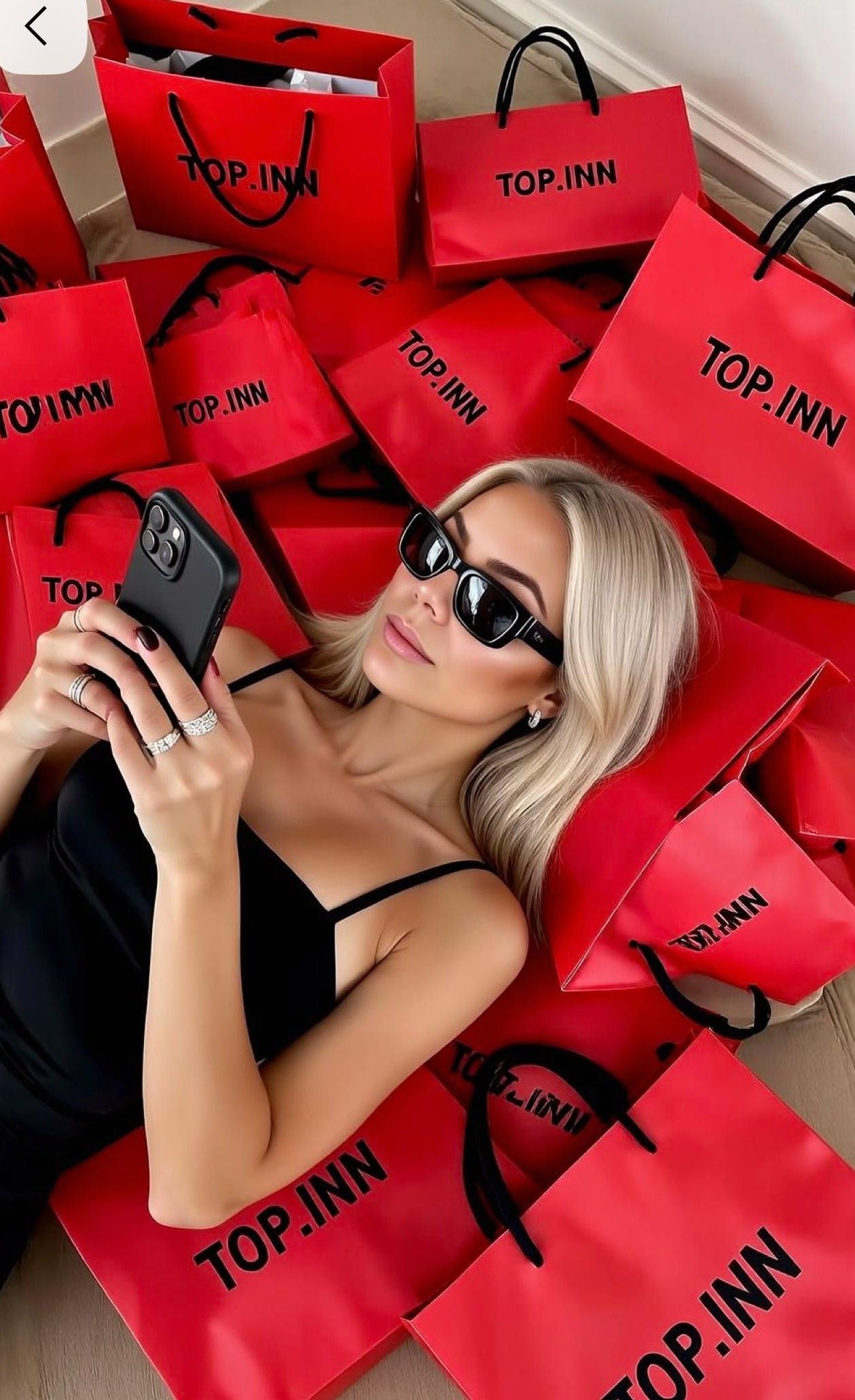 Blonde woman in sunglasses lying among red TOP.INN shopping bags while taking a selfie with her phone.