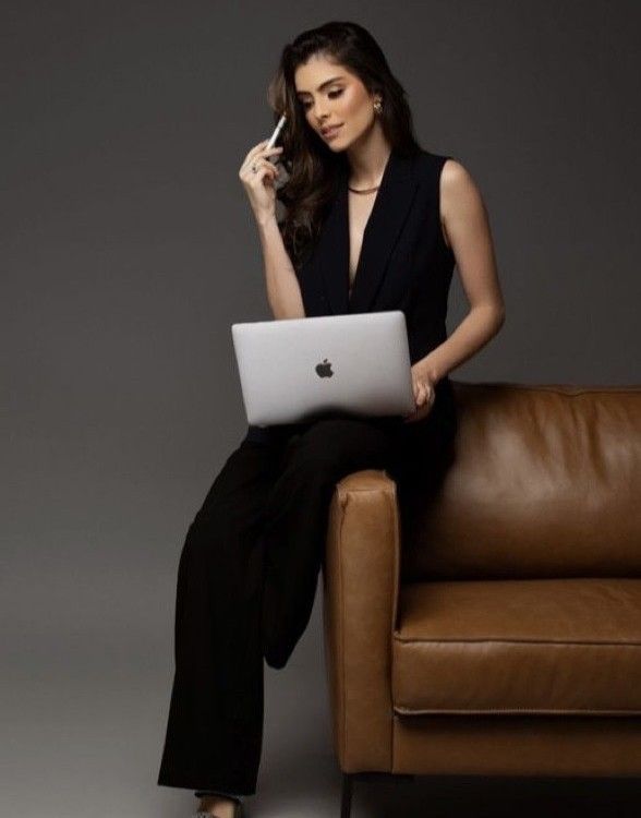 Woman in black jumpsuit sitting on brown leather sofa, holding MacBook laptop and pen, professional business photo.