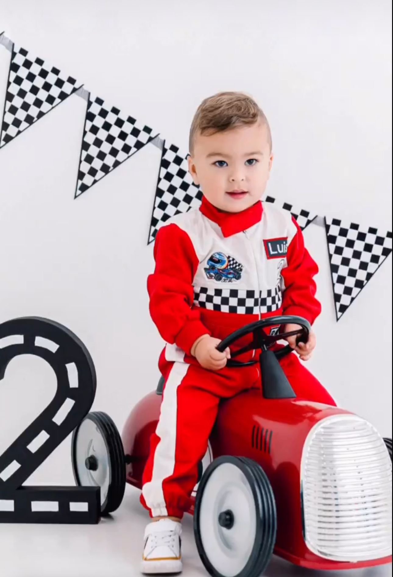 A child in a red and white racing suit sits on a vintage-style toy pedal car with checkered flag decorations in the background.