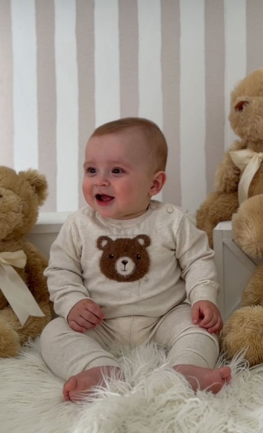 A baby in a cream-colored outfit with teddy bear design lies on white fluffy carpet next to plush bears.