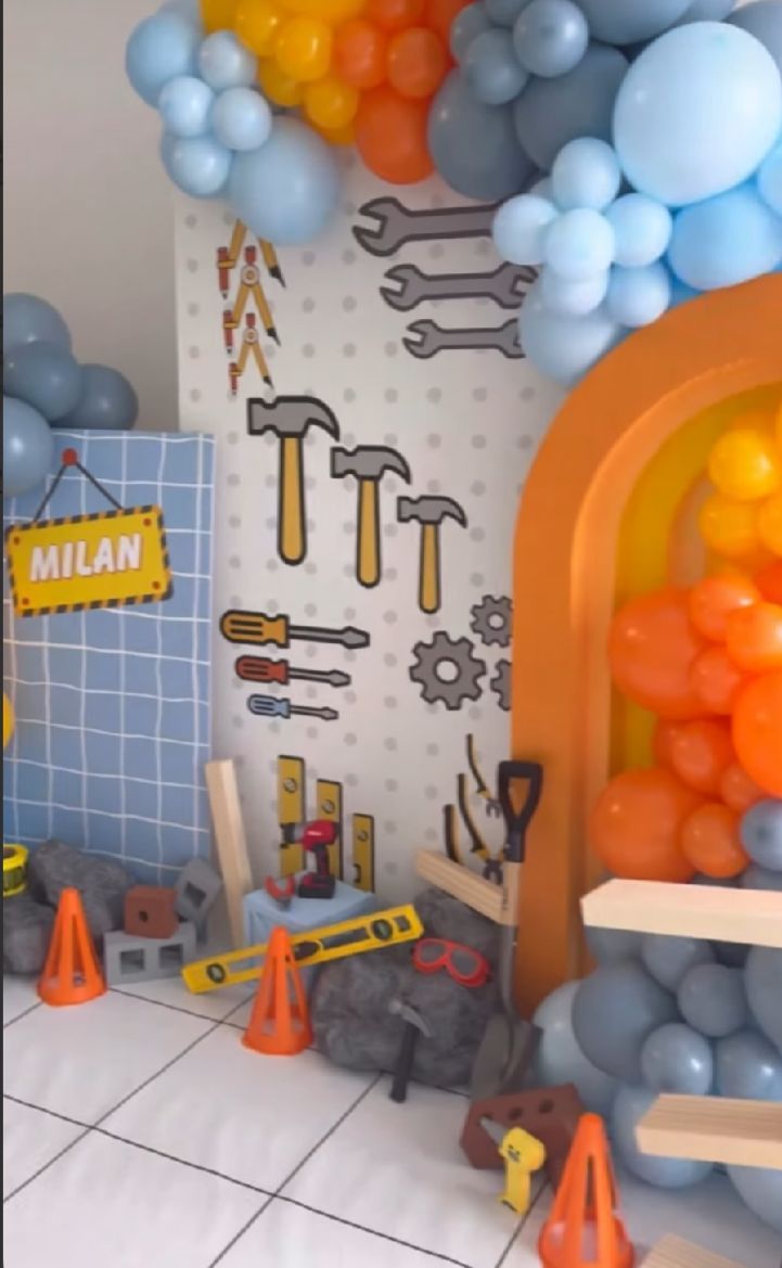 Construction themed party decoration with blue and orange balloons, tool decals, and traffic cones against a white wall.