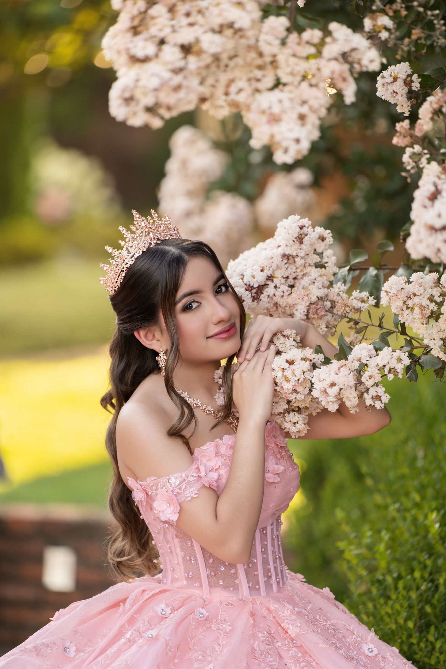 Young woman in pink off-shoulder ball gown with floral crown poses beside blooming white flowers in a garden.