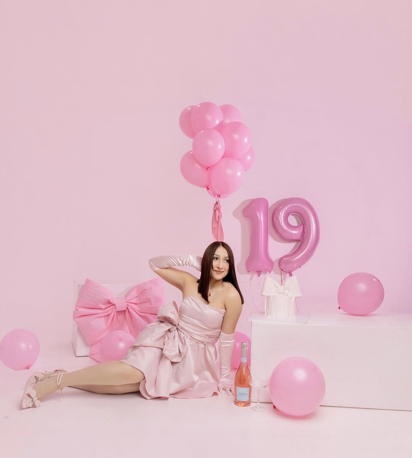 Young woman in pink dress poses with pink balloons and number 19 balloon decorations for a birthday celebration.