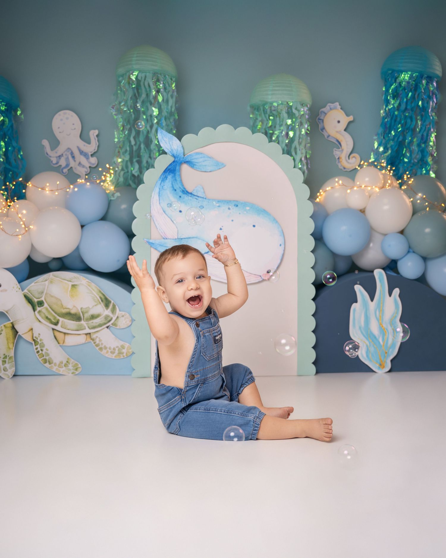Smiling baby boy in denim overalls sits before an ocean-themed backdrop with balloons, sea creatures, and a whale cutout.