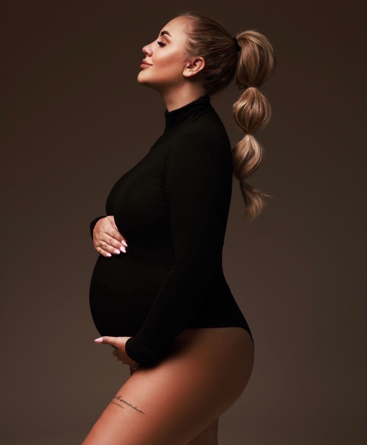 Pregnant woman in black turtleneck bodysuit cradling baby bump, side profile pose against dark brown background.