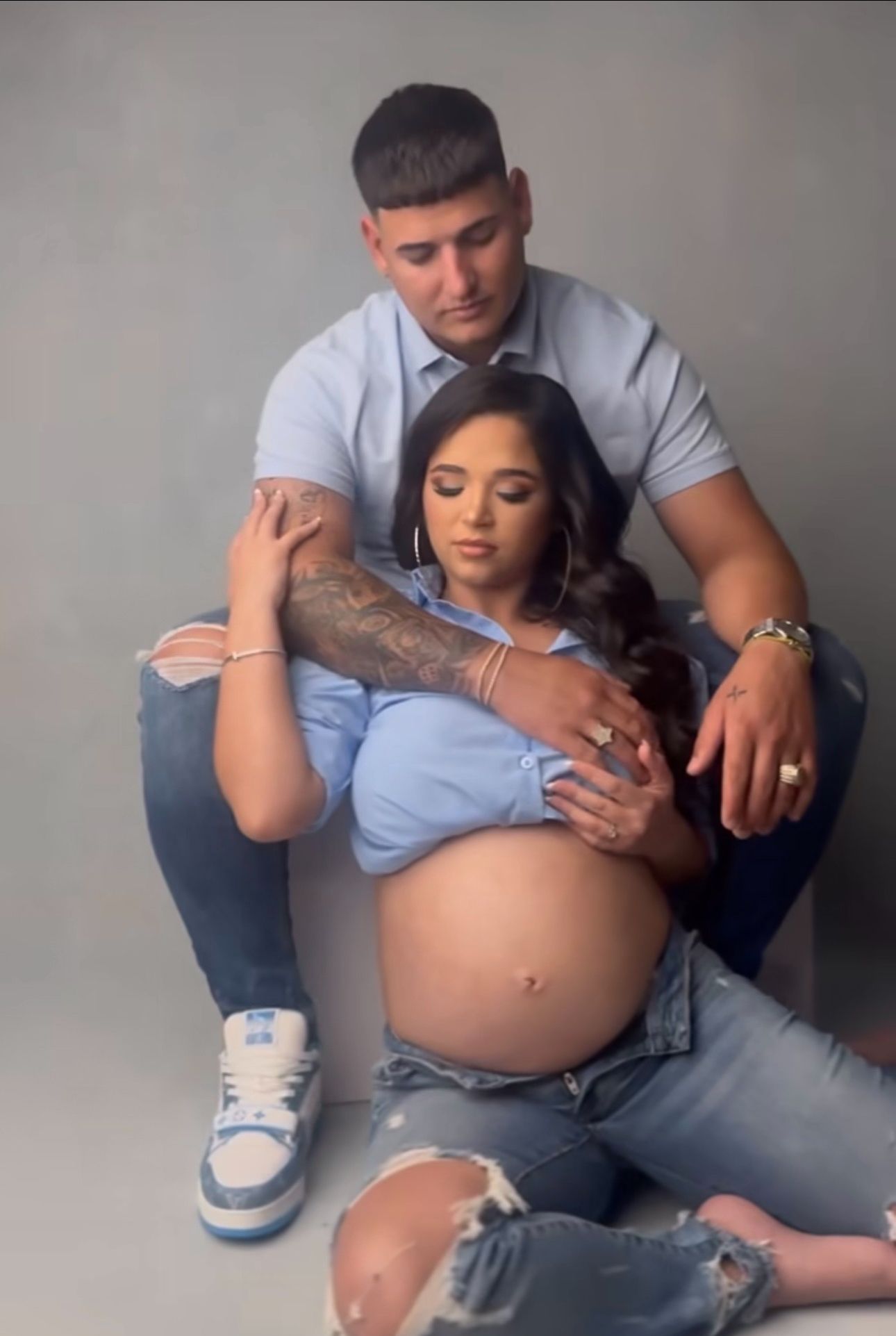 Couple in blue outfits poses for maternity photo shoot, woman showing baby bump, man sitting behind her.