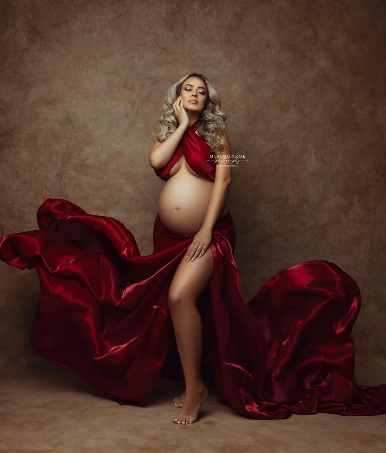 Pregnant woman posing in flowing red silk fabric for a maternity photography session against a brown studio backdrop.