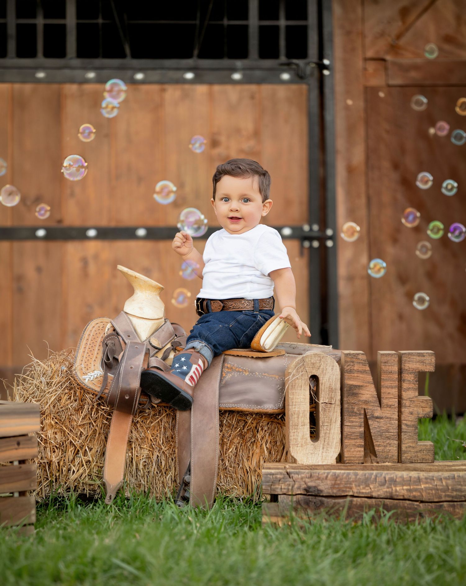 Toddler boy sitting on hay bale with wooden ONE sign, cowboy boots, saddle, and bubbles floating near barn doors.