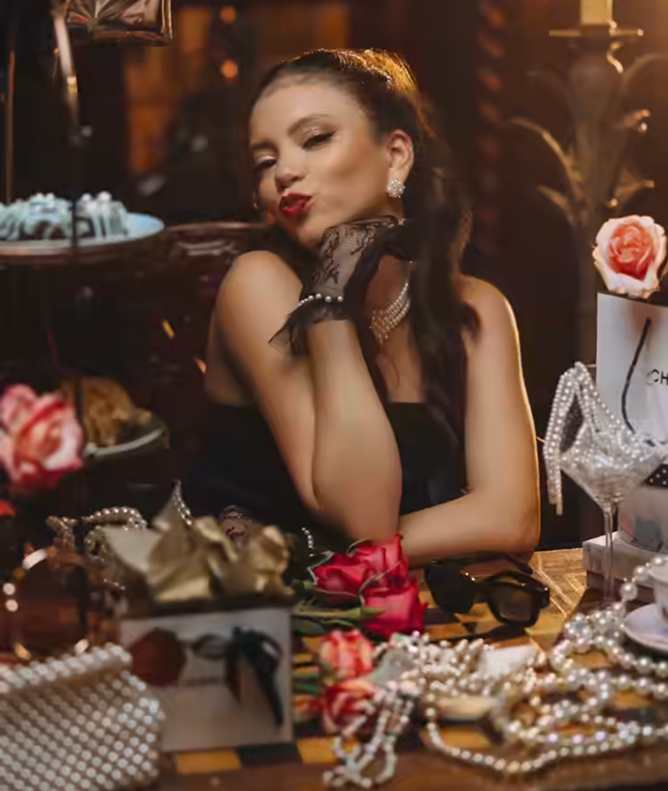 A glamorous woman in black poses at a table adorned with roses, pearls, and jewelry in a luxurious setting.