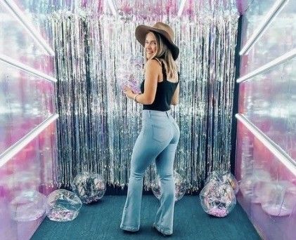 Woman in flare jeans, black top, and tan hat posing in a shimmery silver tinsel photo booth.
