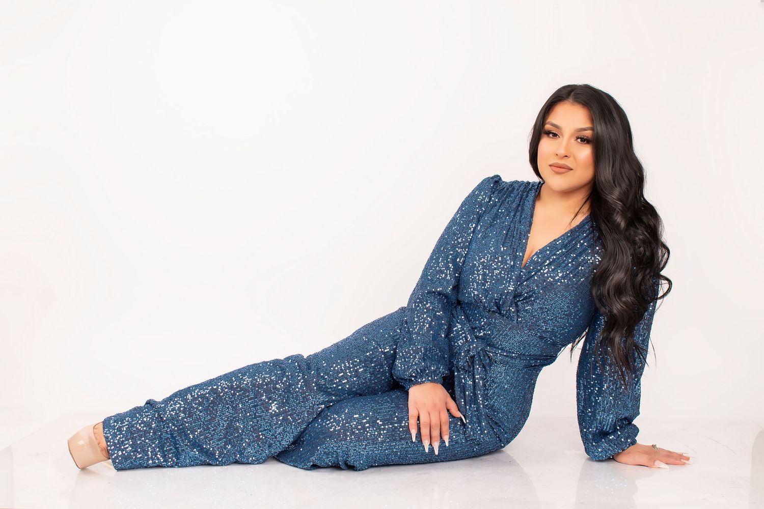 Woman with long wavy dark hair reclining on a white floor in a sparkling blue sequin jumpsuit with a wrap-style top.