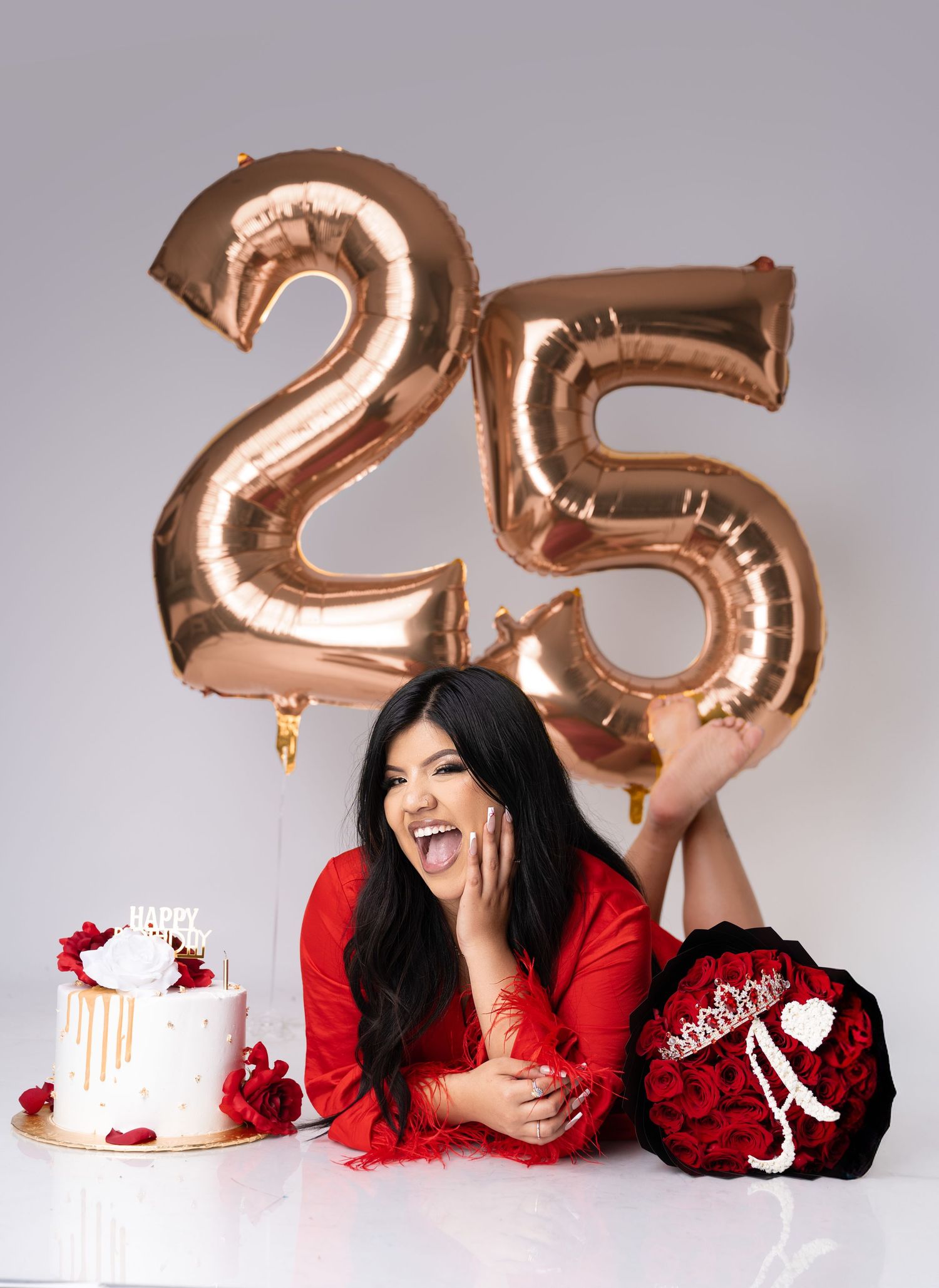 Woman in a red outfit laughs while posing beside rose gold 25 balloons, a white birthday cake, and a red rose bouquet with a tiara.