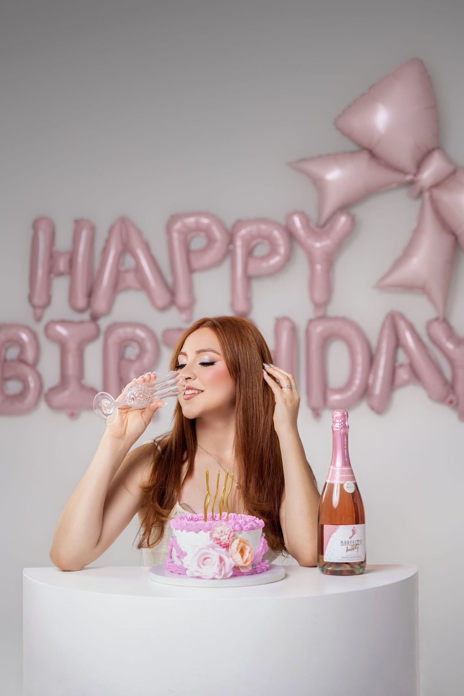 Woman with long red hair sipping champagne beside a pink flower cake and rosé bottle with pink Happy Birthday balloons behind her.