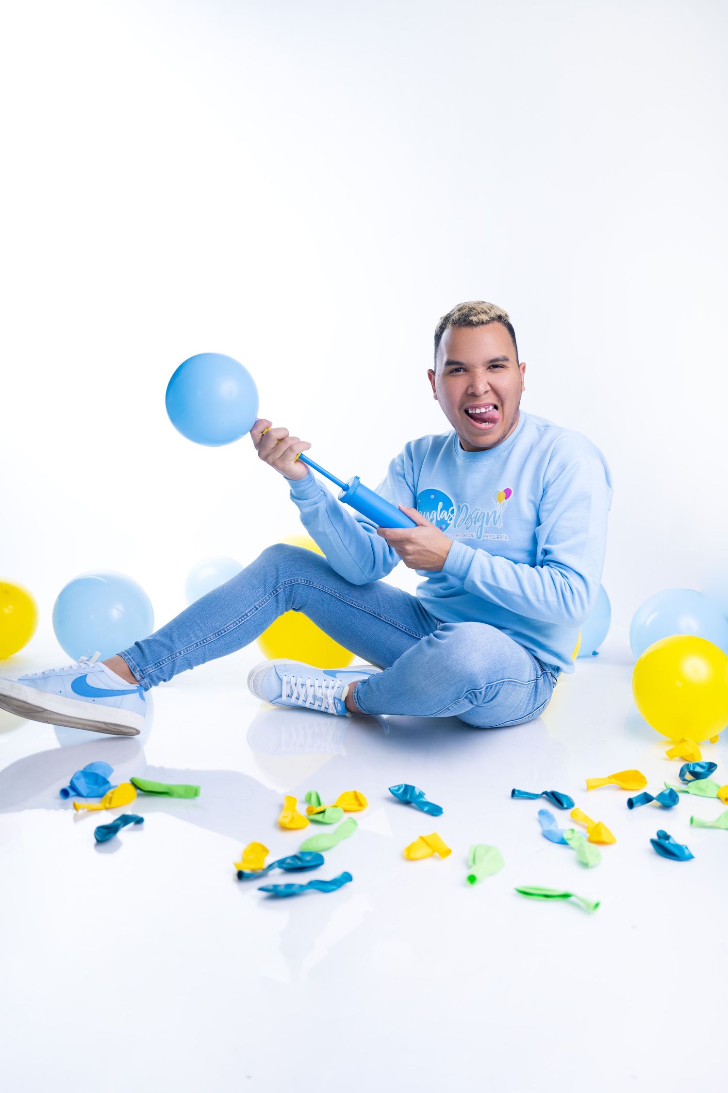 Man in blue sweatshirt sitting on white floor with colorful balloons, holding balloon pump, smiling playfully.