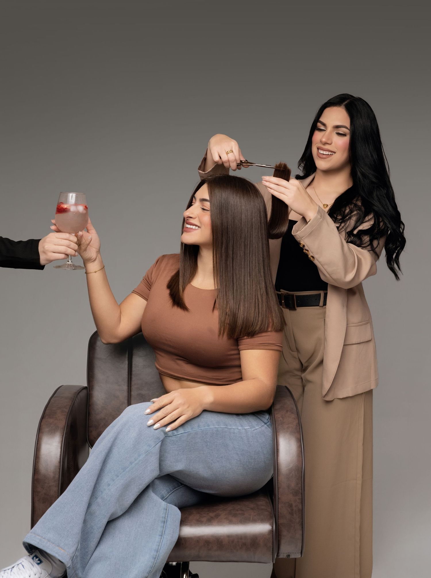 Woman sitting in salon chair getting hair styled while holding a cocktail, with a smiling stylist working behind her.