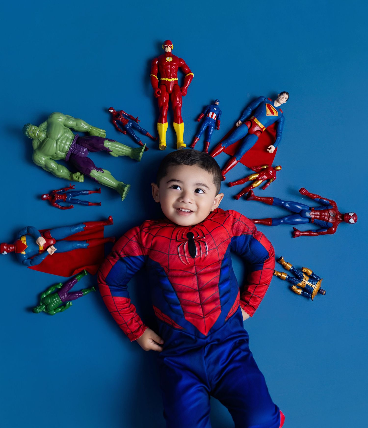 Young boy in Spider-Man costume lying on blue floor surrounded by superhero action figures including Hulk and Flash.