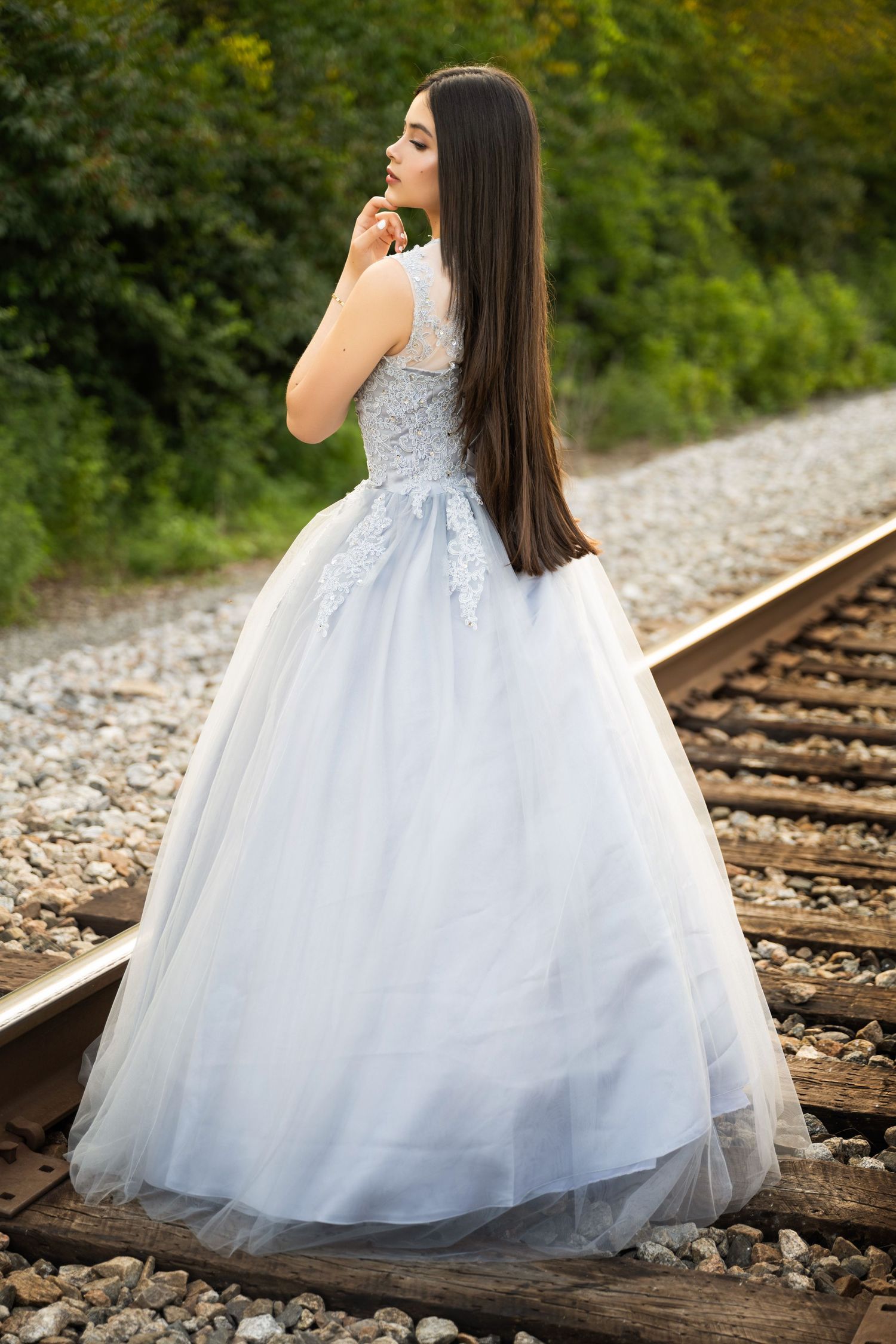 Young woman in white ball gown with lace bodice stands on railway tracks surrounded by lush greenery.