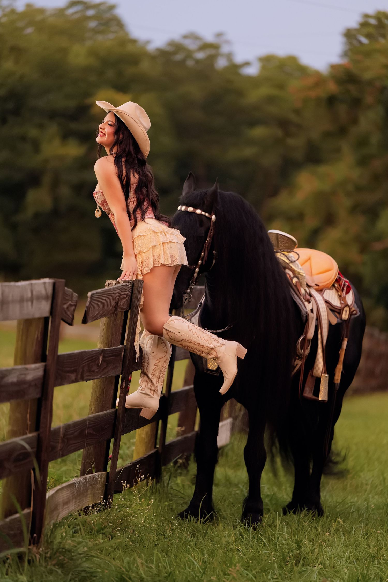A woman in cowgirl attire with boots and hat leans on a wooden fence beside a black horse in a green pasture.