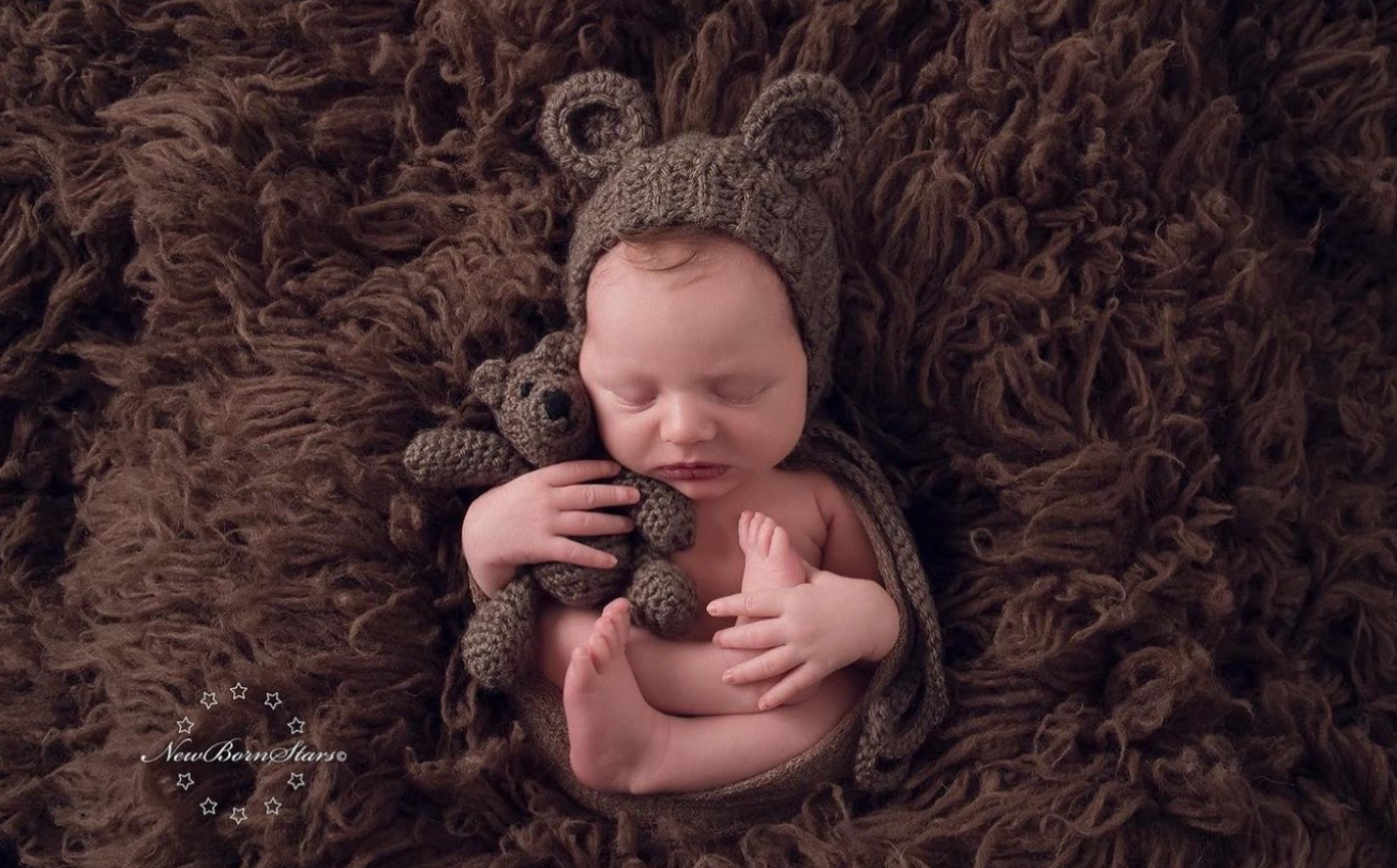 Sleeping newborn wearing knitted brown bear bonnet lying on brown textured blanket with teddy bear.