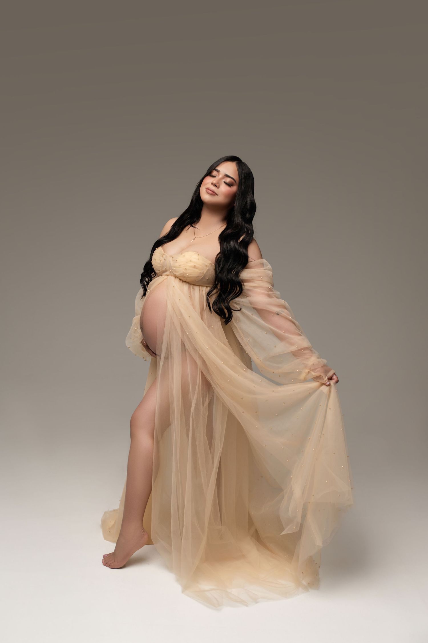A pregnant woman in a flowing champagne-colored tulle gown poses gracefully against a neutral studio backdrop.