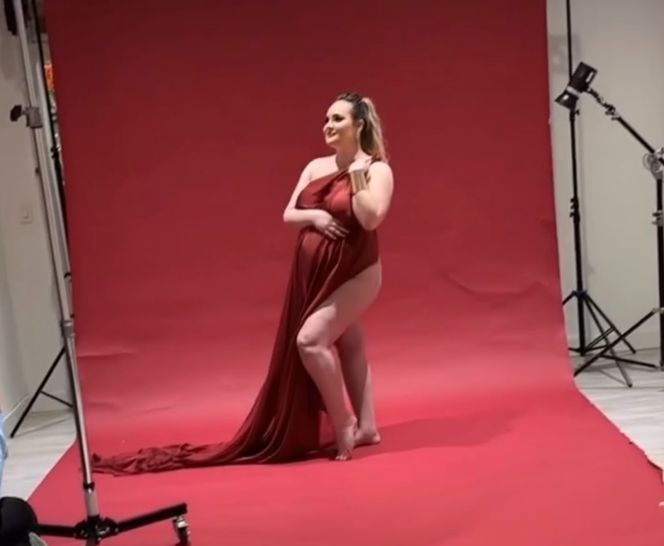 Professional maternity photo shoot showing a woman in a red dress posing in a studio with pink backdrop lighting.